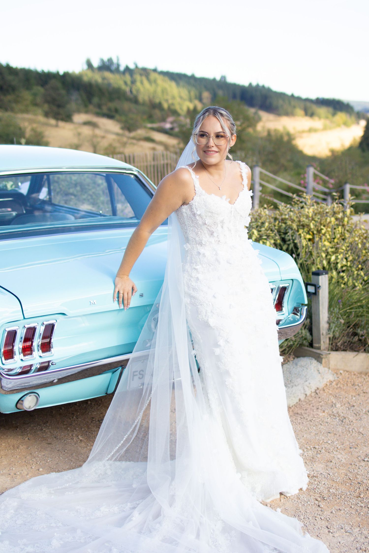 Bride in white floral gown with veil poses beside a classic light blue Ford Mustang outdoors.