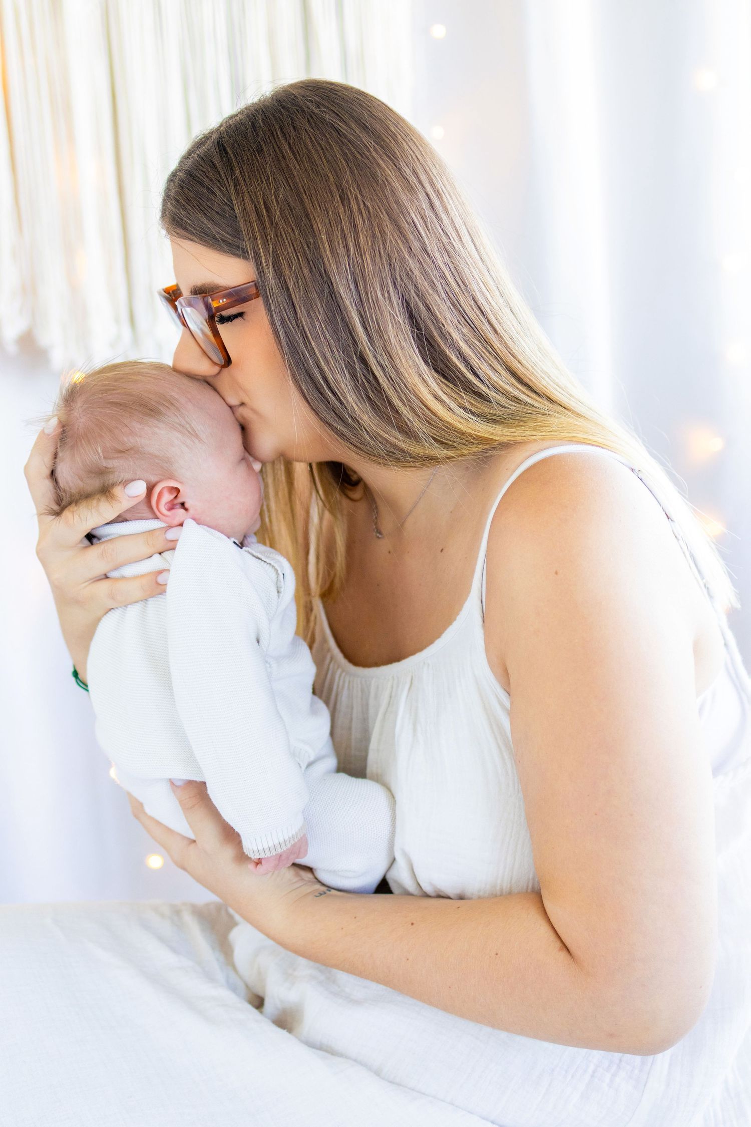 A young mother with long hair and glasses gently kisses her newborn baby while sitting on a bed.