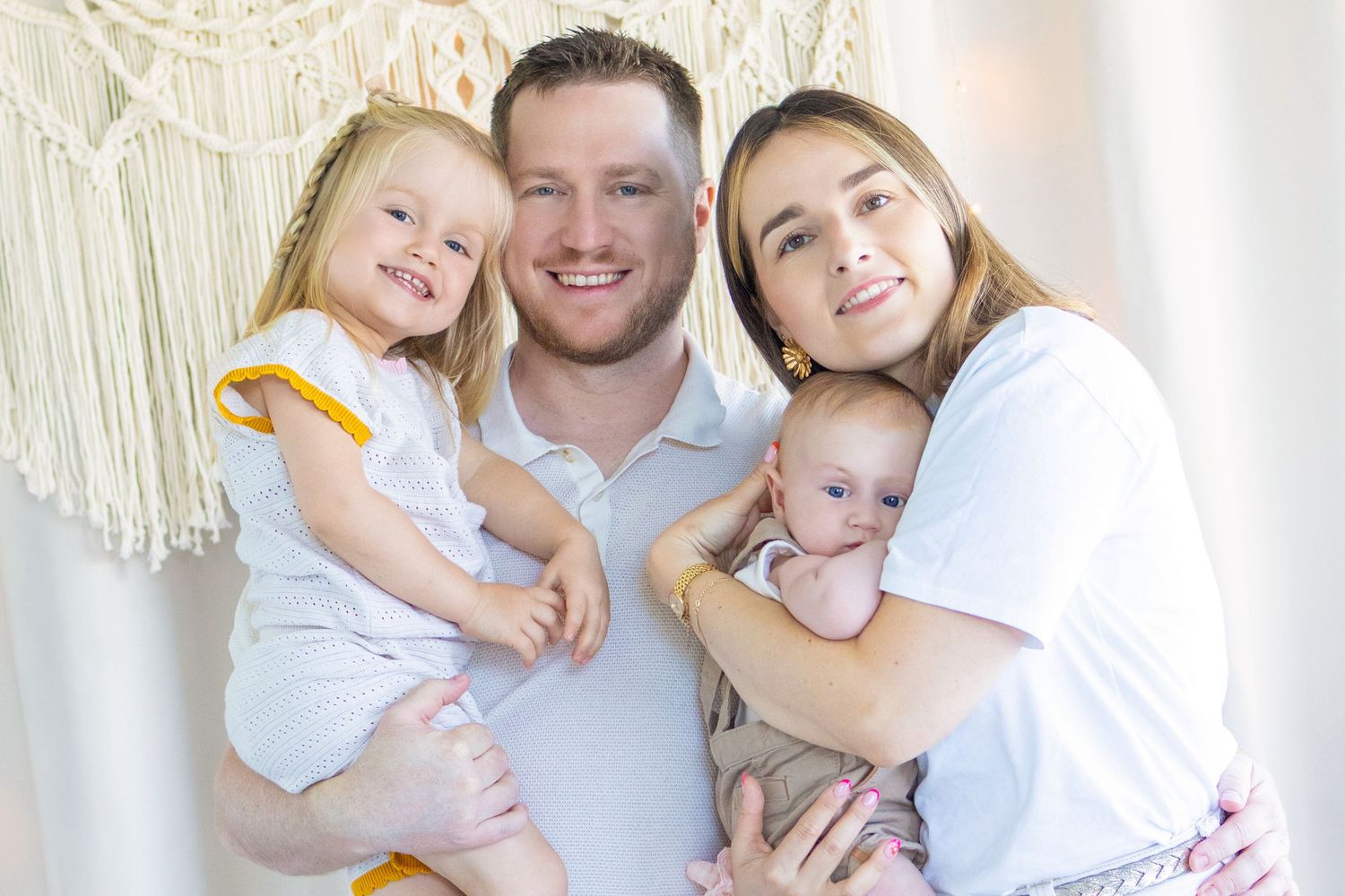 Happy family of four posing together, parents holding a toddler girl and newborn baby in a bright, cozy room.