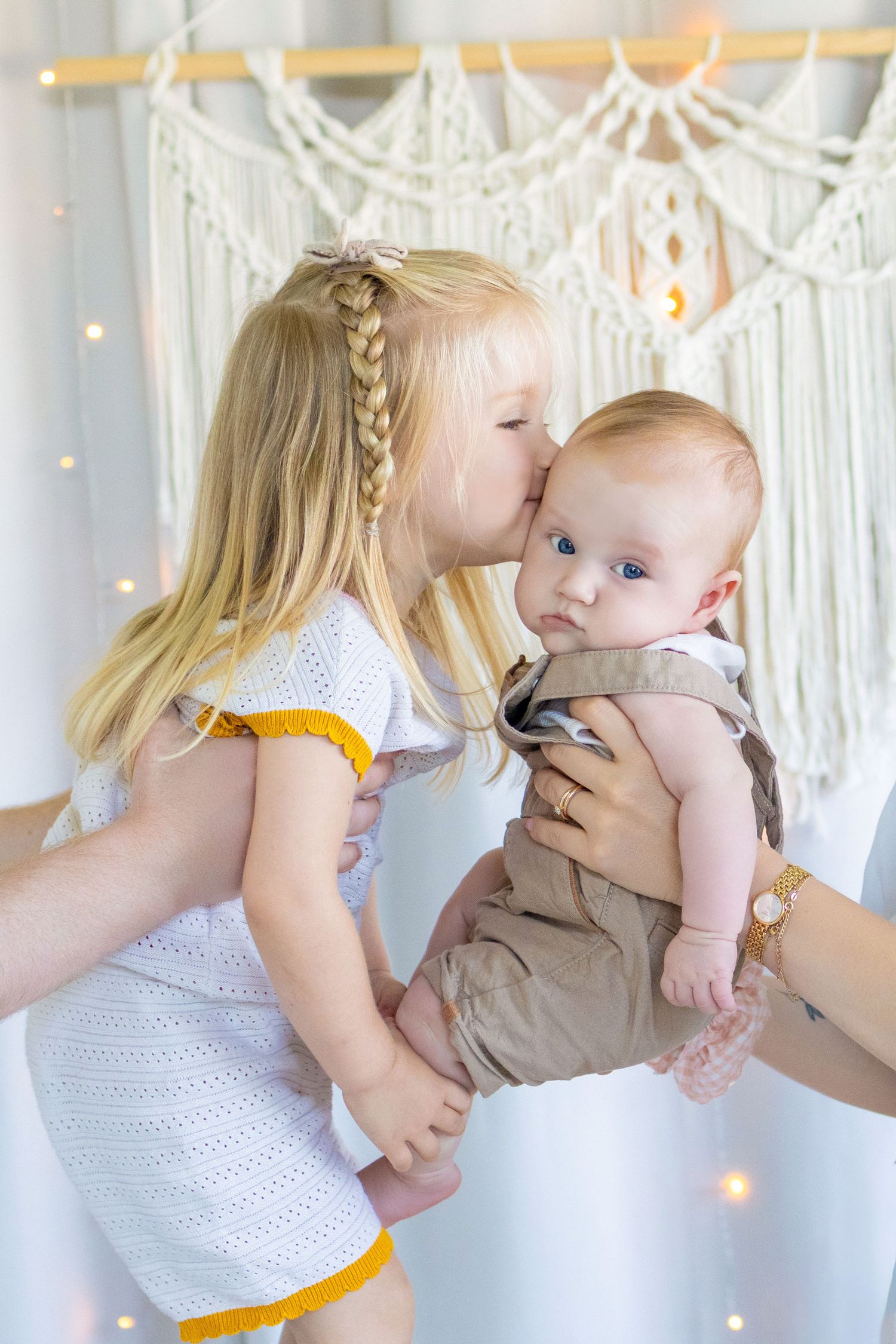 Blonde girl in white dress kissing baby brother held by adult hands, with macrame wall decor and fairy lights.