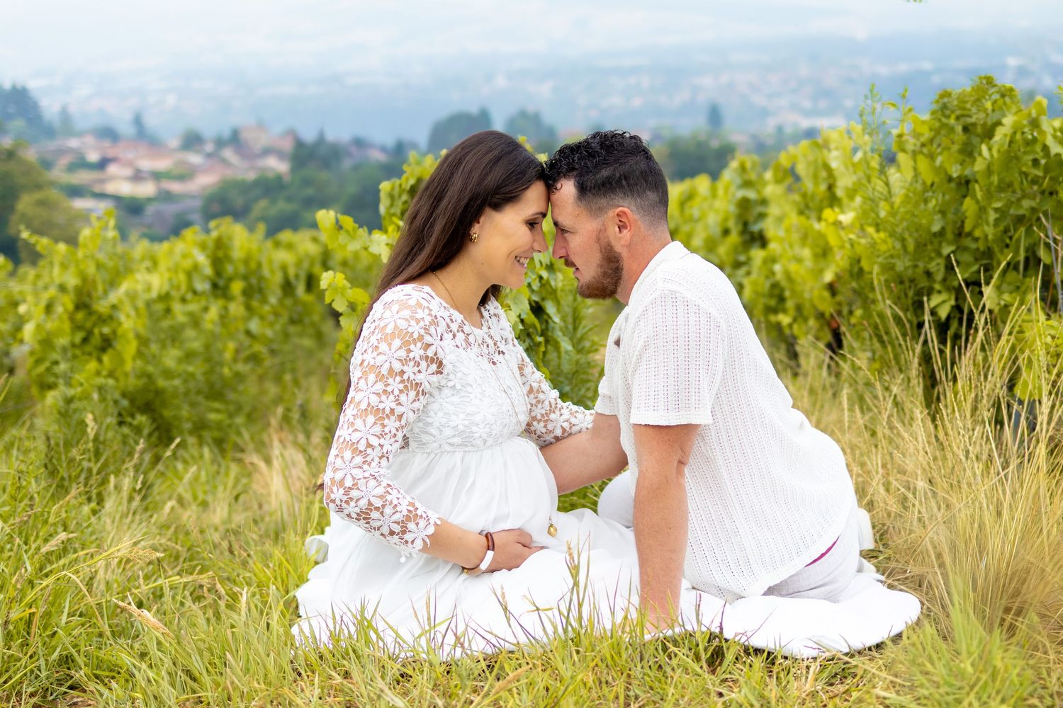 Pregnant couple in white sitting in lush green field, sharing tender moment with hands on baby bump, vineyard view behind.