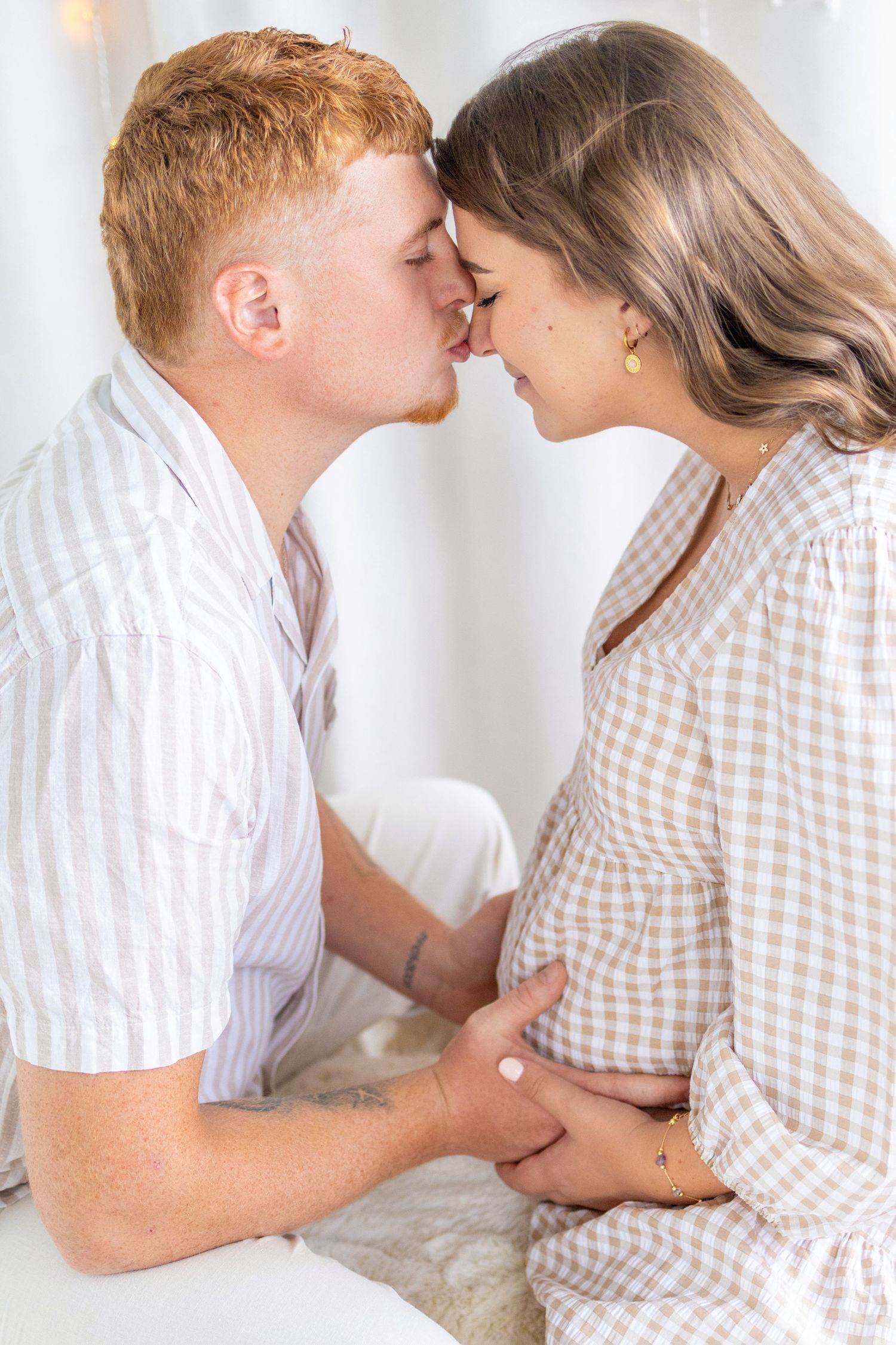 Couple sharing a tender kiss, pregnant woman in gingham dress with partner in striped shirt in bright white room.