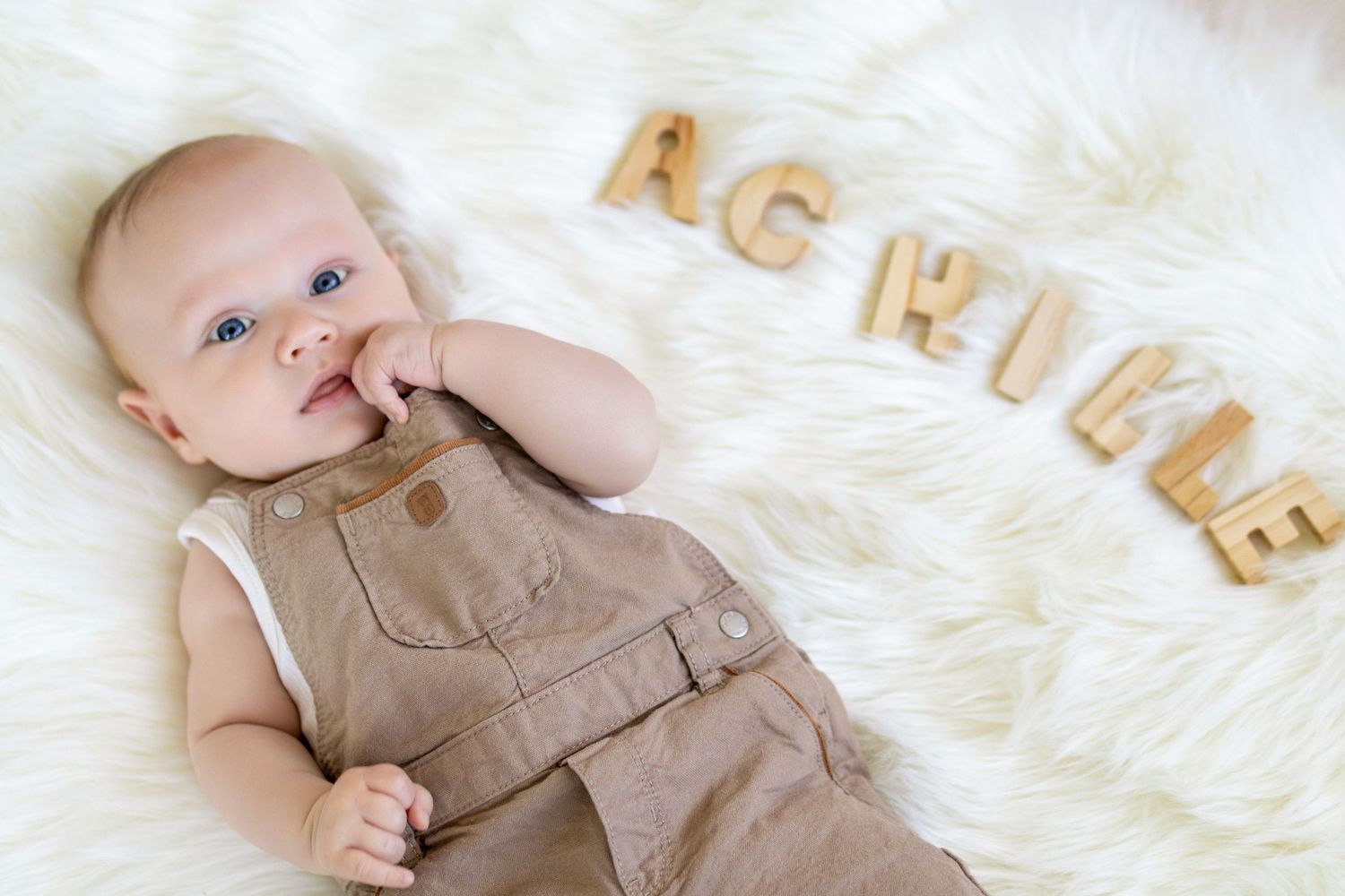 A cute baby boy in brown overalls lies on a fluffy white rug next to wooden letters spelling Achille.