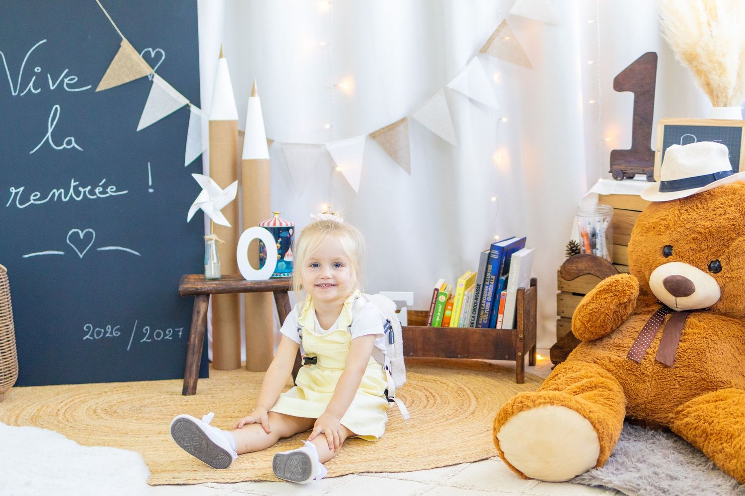 Toddler girl sitting on a round rug in a decorated playroom with a giant teddy bear and a chalkboard sign.