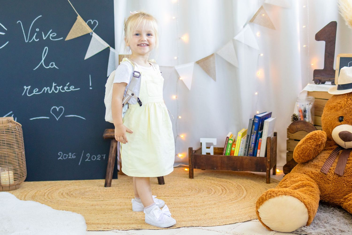 Smiling toddler girl in yellow dress stands by chalkboard with back-to-school decor, teddy bear, and fairy lights.