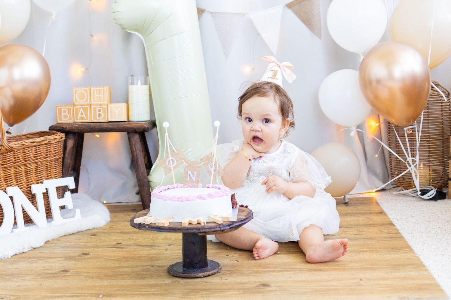 Baby girl in white dress sits beside a pink first birthday cake surrounded by gold and white balloons and decorations.