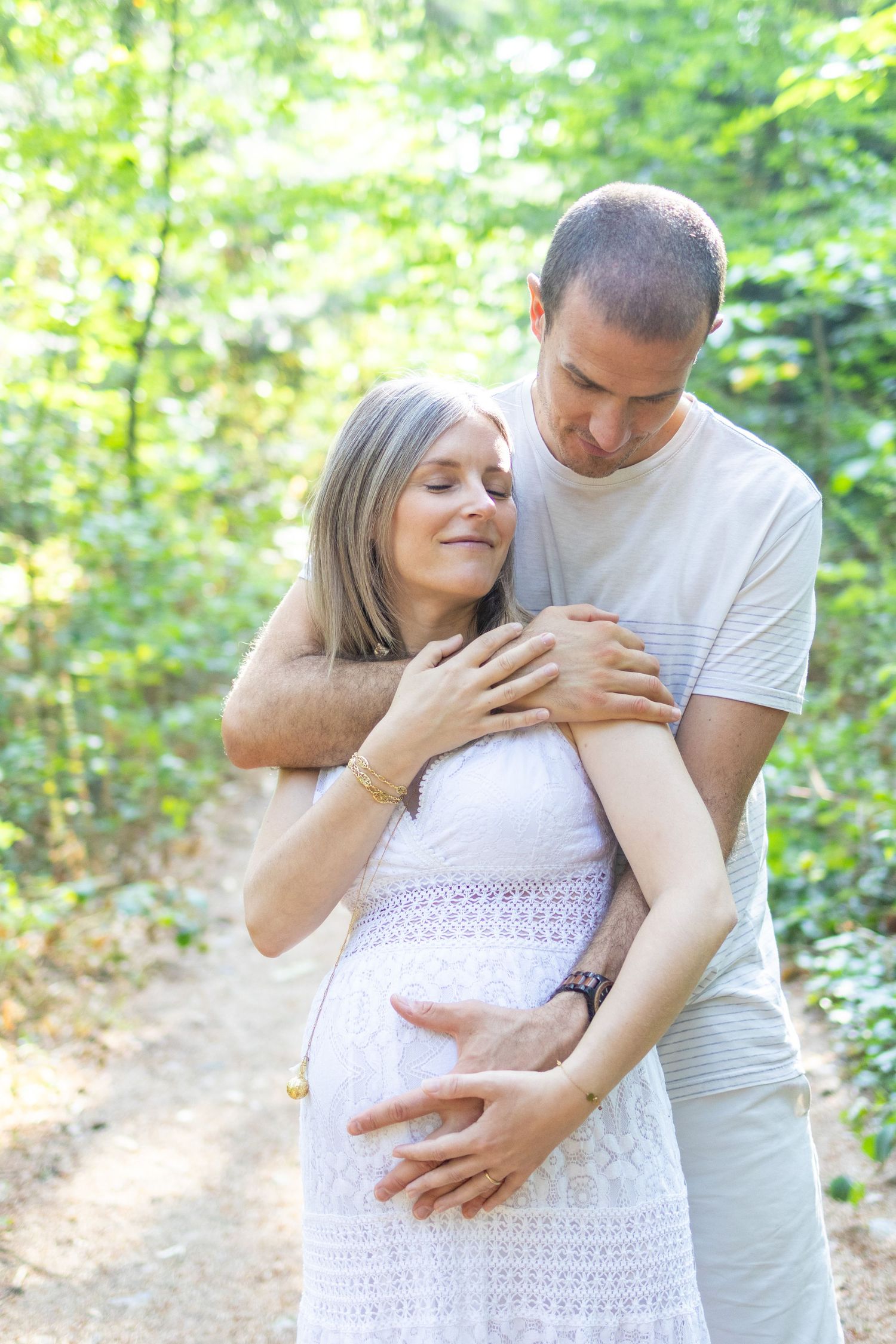 Couple in a forest, man embracing pregnant woman from behind, both smiling peacefully on a sunlit path.