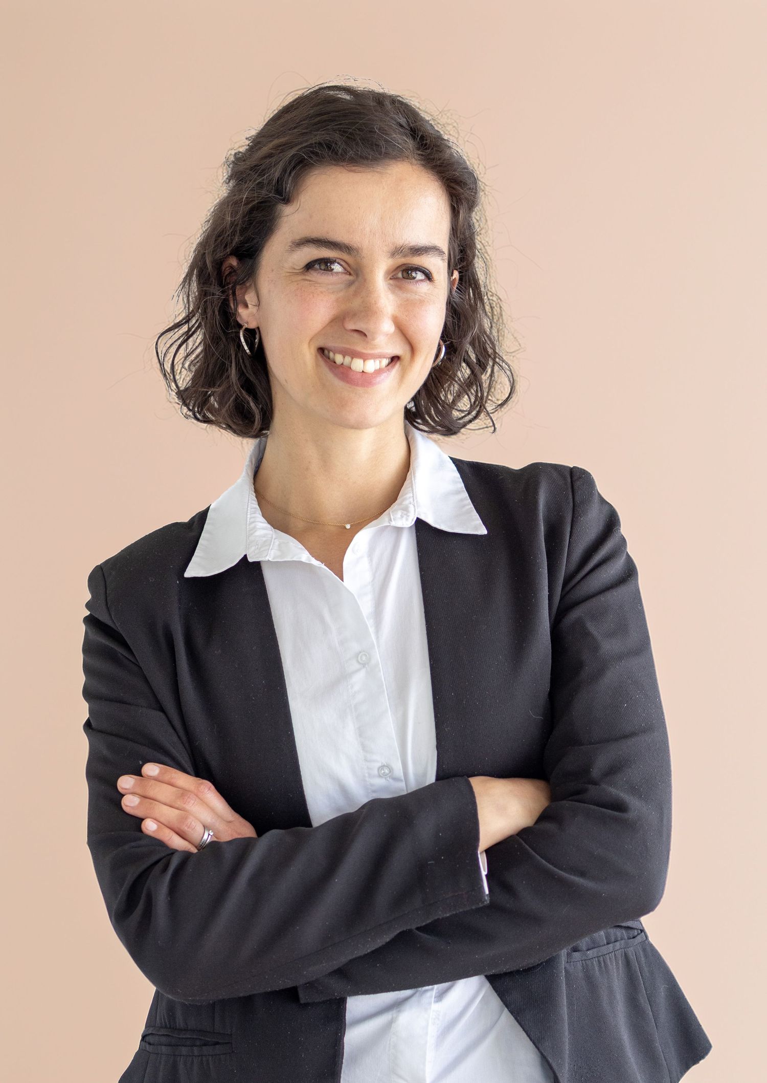 Professional businesswoman in black blazer and white shirt smiling with arms crossed against beige background.