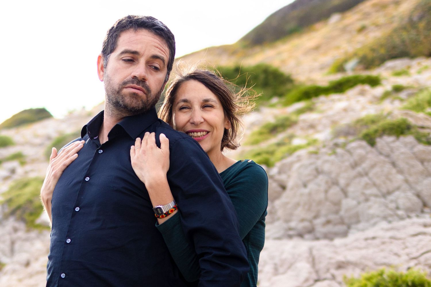 Couple embracing outdoors near rocky cliffs, woman smiling over man's shoulder in a coastal setting.