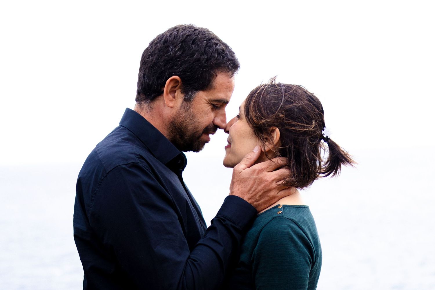 A couple shares an intimate moment, nearly kissing outdoors by the ocean on an overcast day.