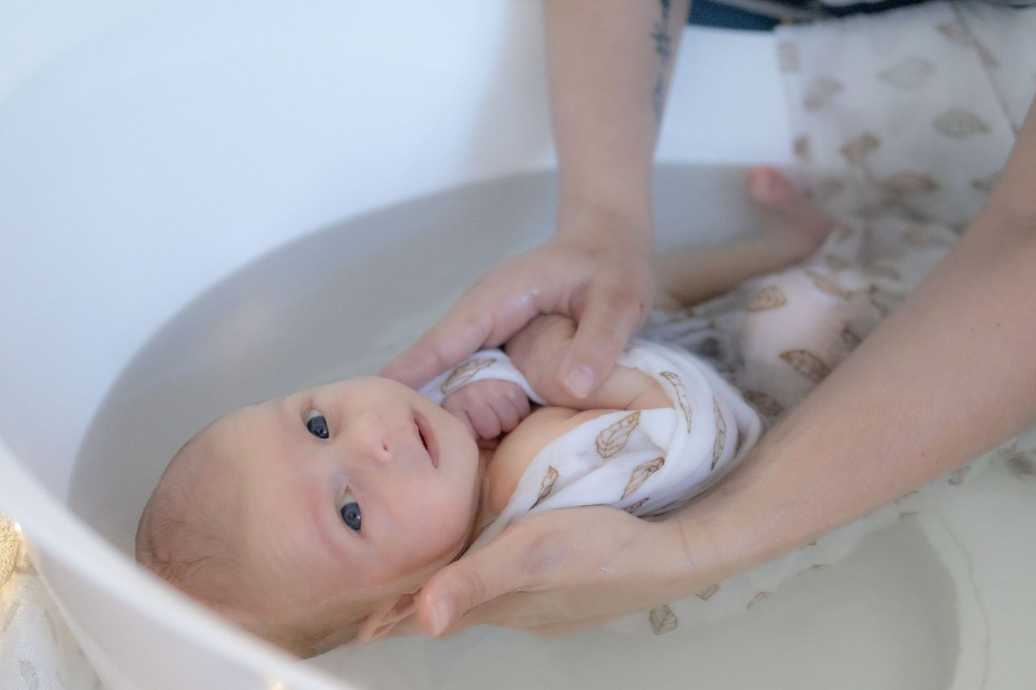 Newborn baby in a white feather-print outfit being gently bathed in a round white baby tub.