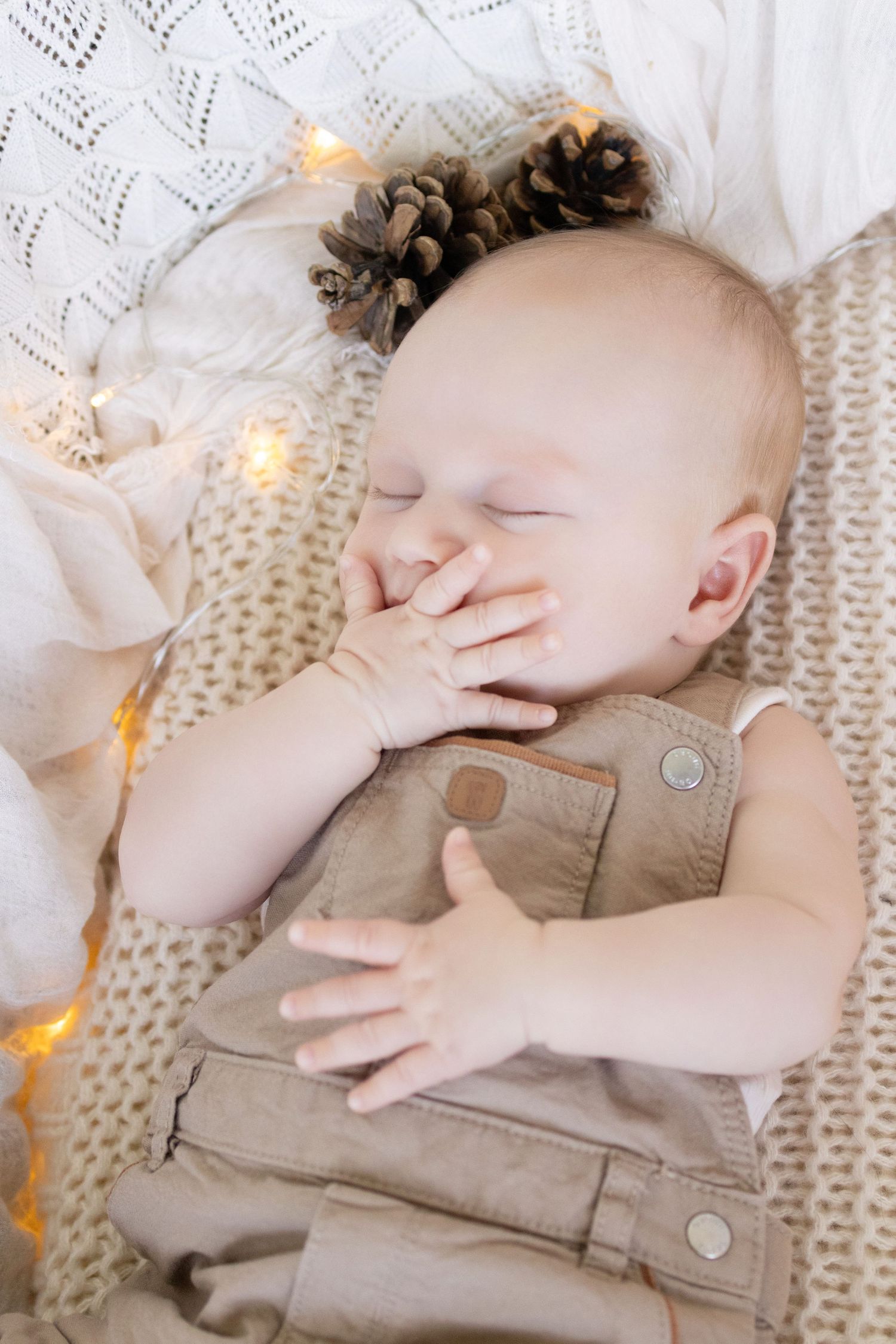 Sleeping baby in tan overalls on a knit blanket with pine cones and warm fairy lights.