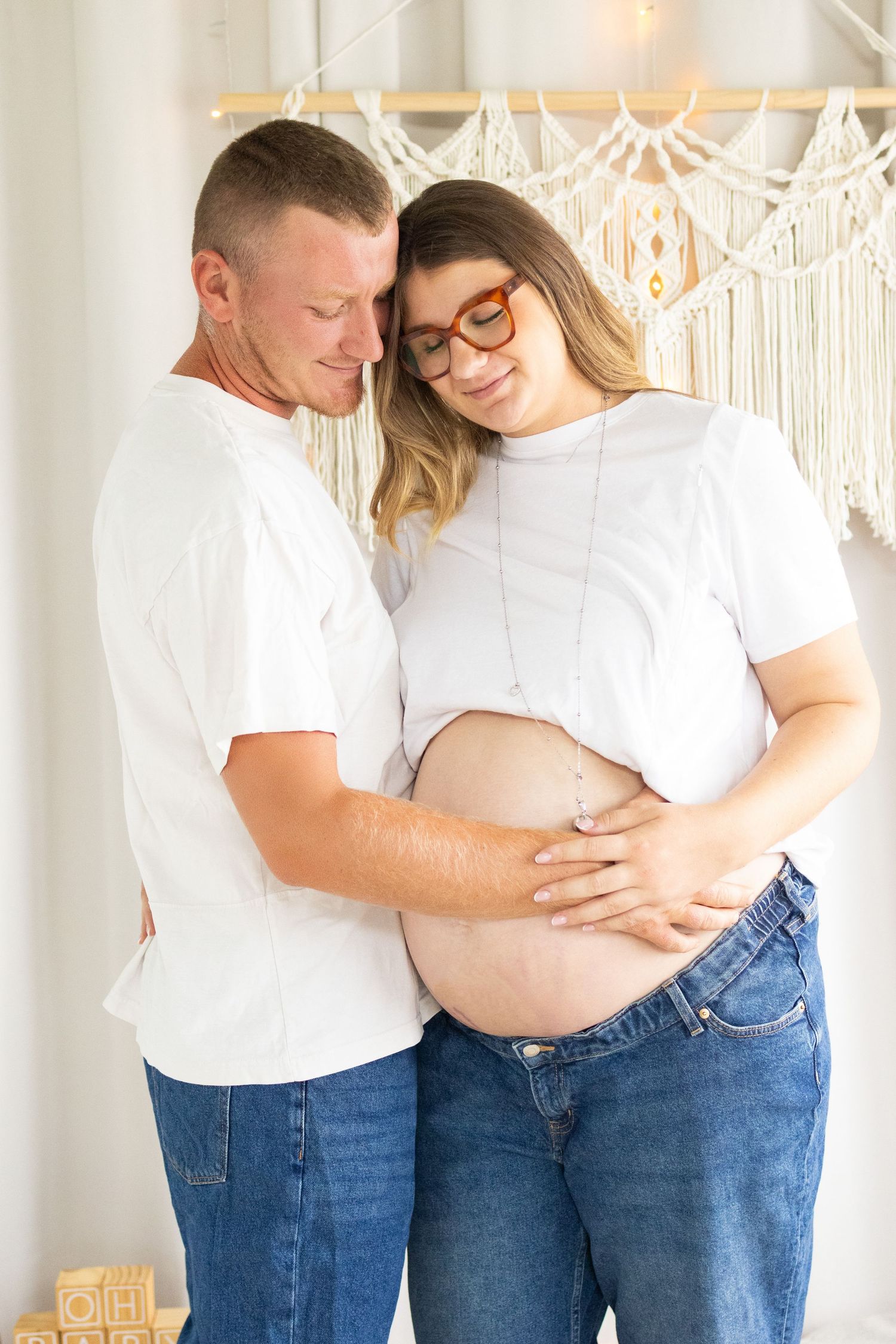 Couple in white shirts and jeans tenderly cradling pregnant belly during maternity photo session.