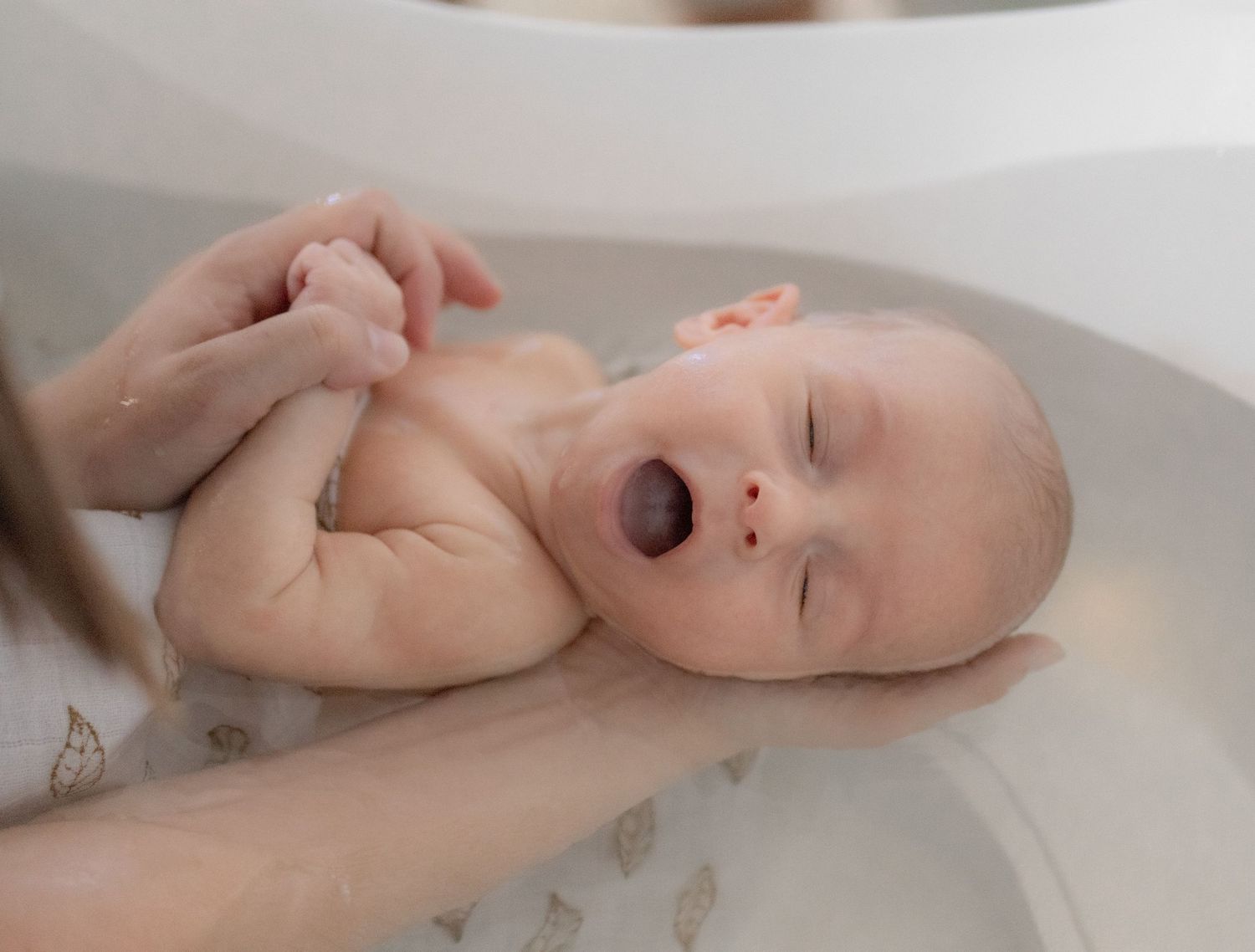 Newborn baby yawning during a gentle bath, supported by caring hands in a white baby bathtub.