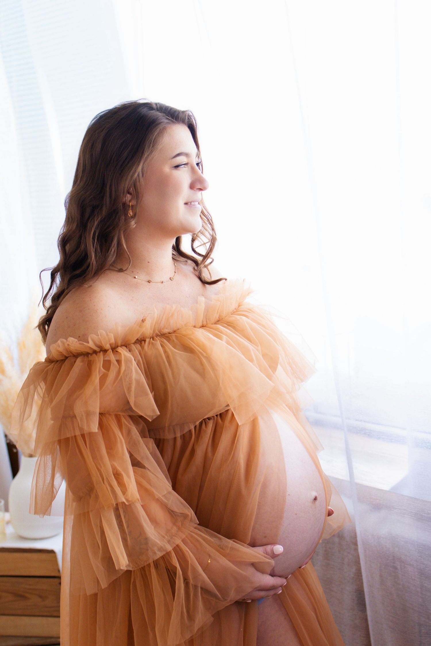 Pregnant woman in elegant peach tulle off-shoulder gown cradling baby bump, smiling softly near bright window.