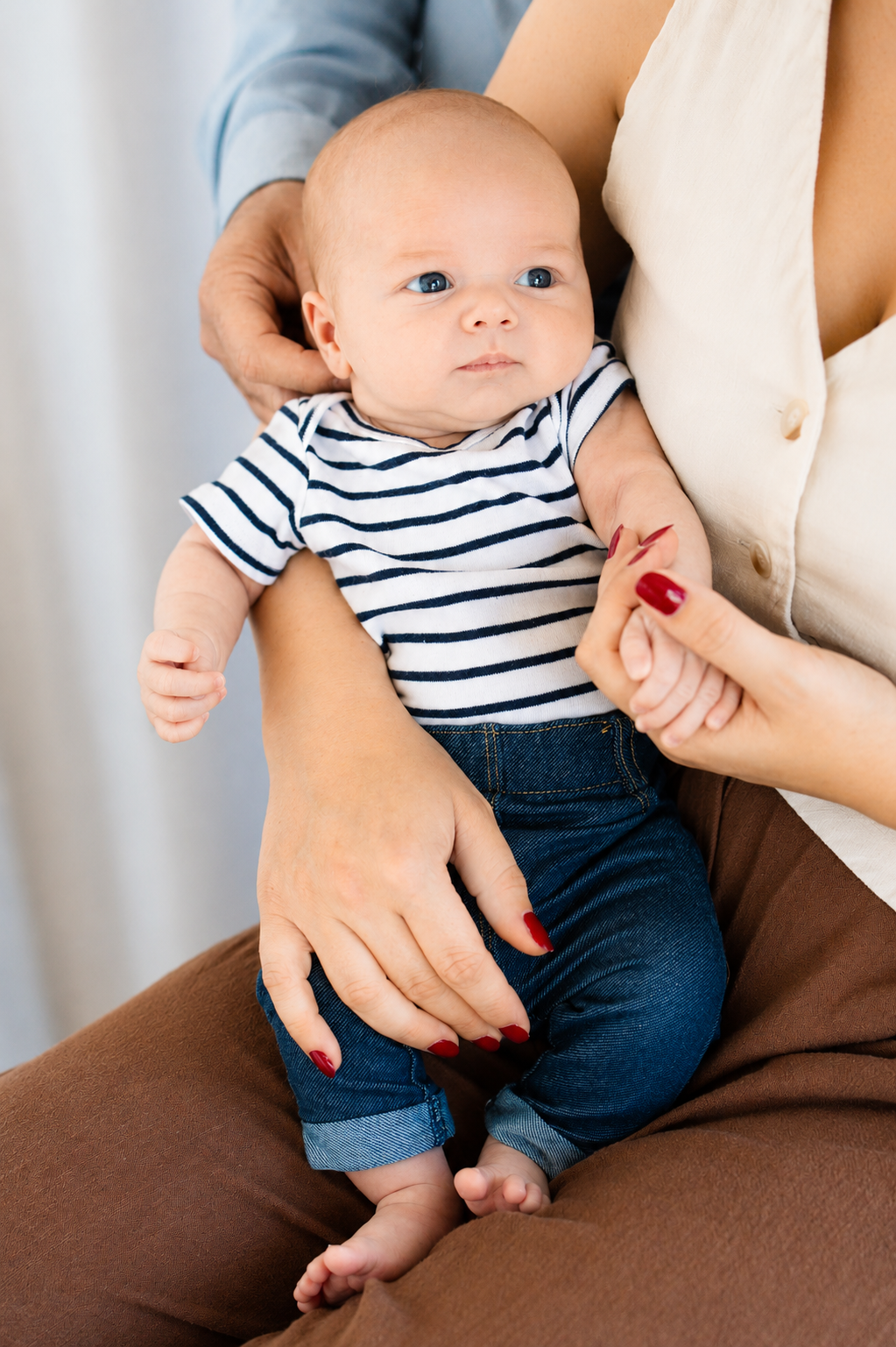 A baby in a striped onesie and jeans is held by two adults with red nails in a bright, cozy setting.