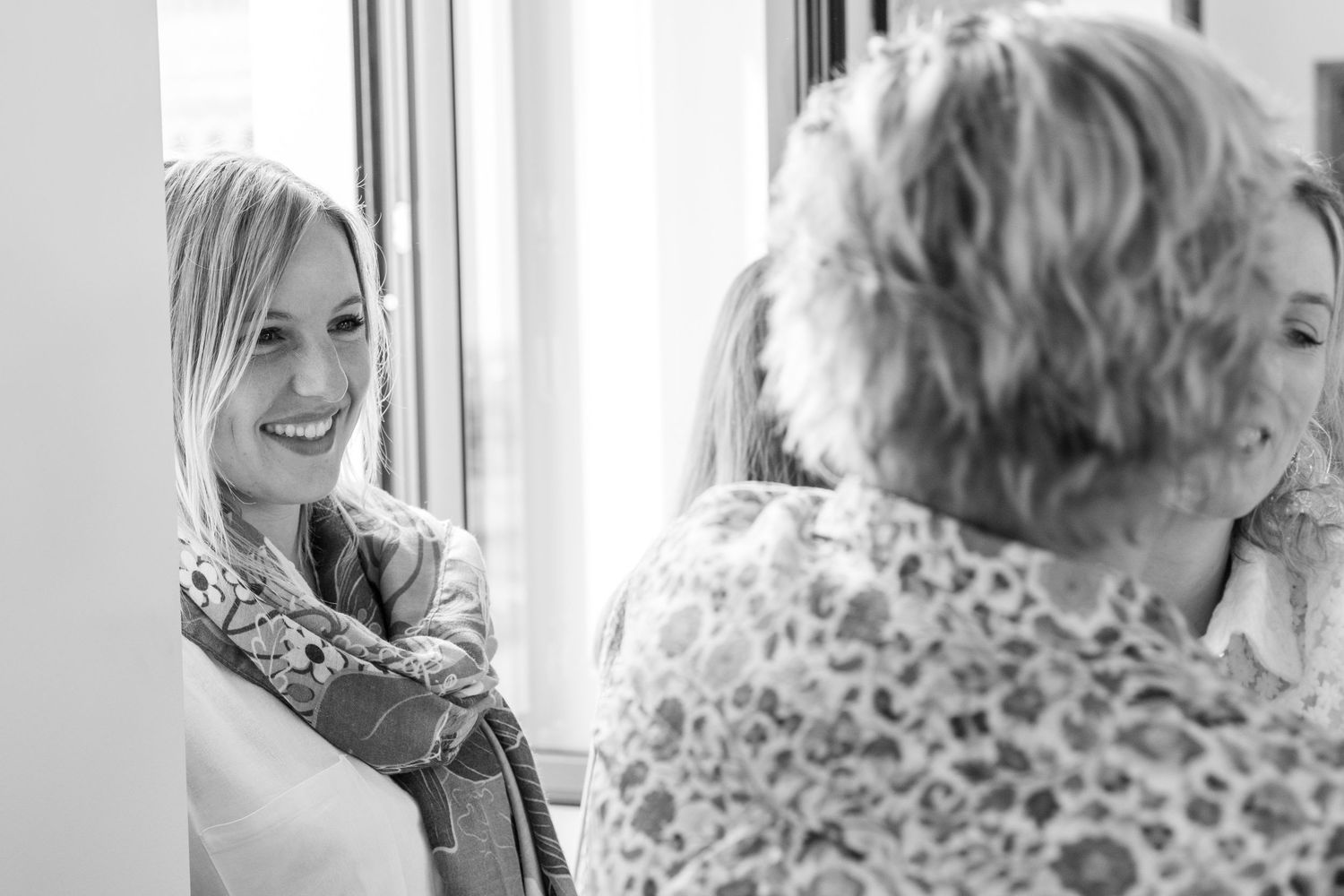 Black and white photo of smiling blonde woman with scarf talking to older woman near bright window.