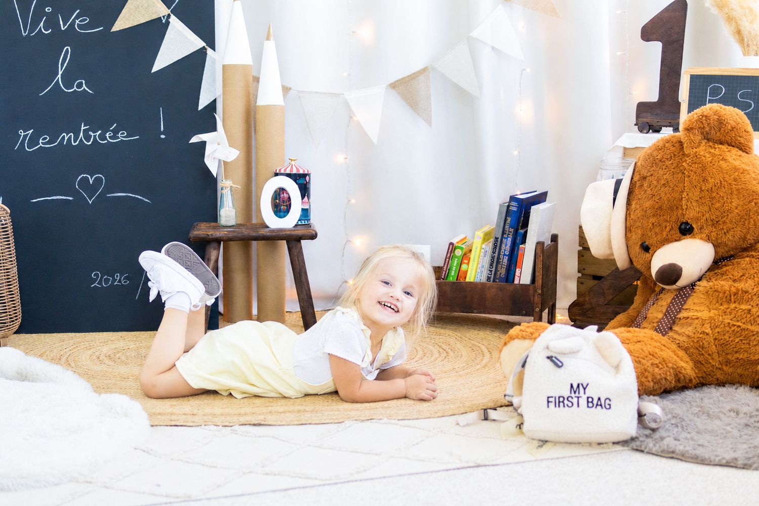 Smiling blonde girl lying on floor in playroom with large teddy bear, chalkboard wall, and colorful books.