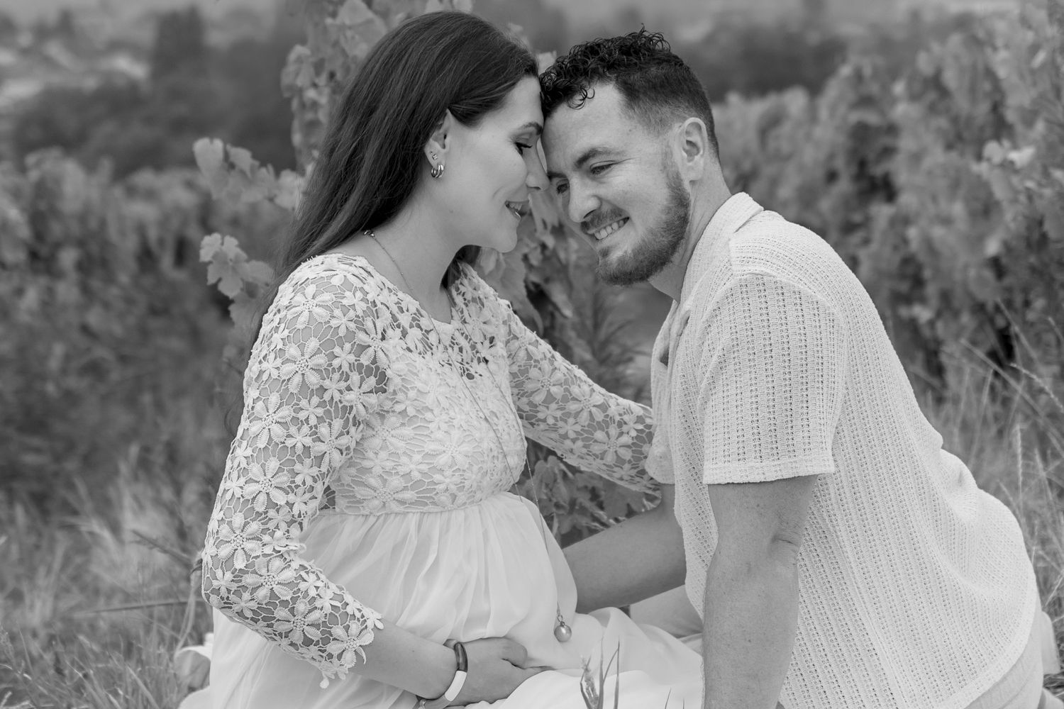 Black and white maternity photo of a couple sitting outdoors, smiling at each other while cradling a pregnant belly.