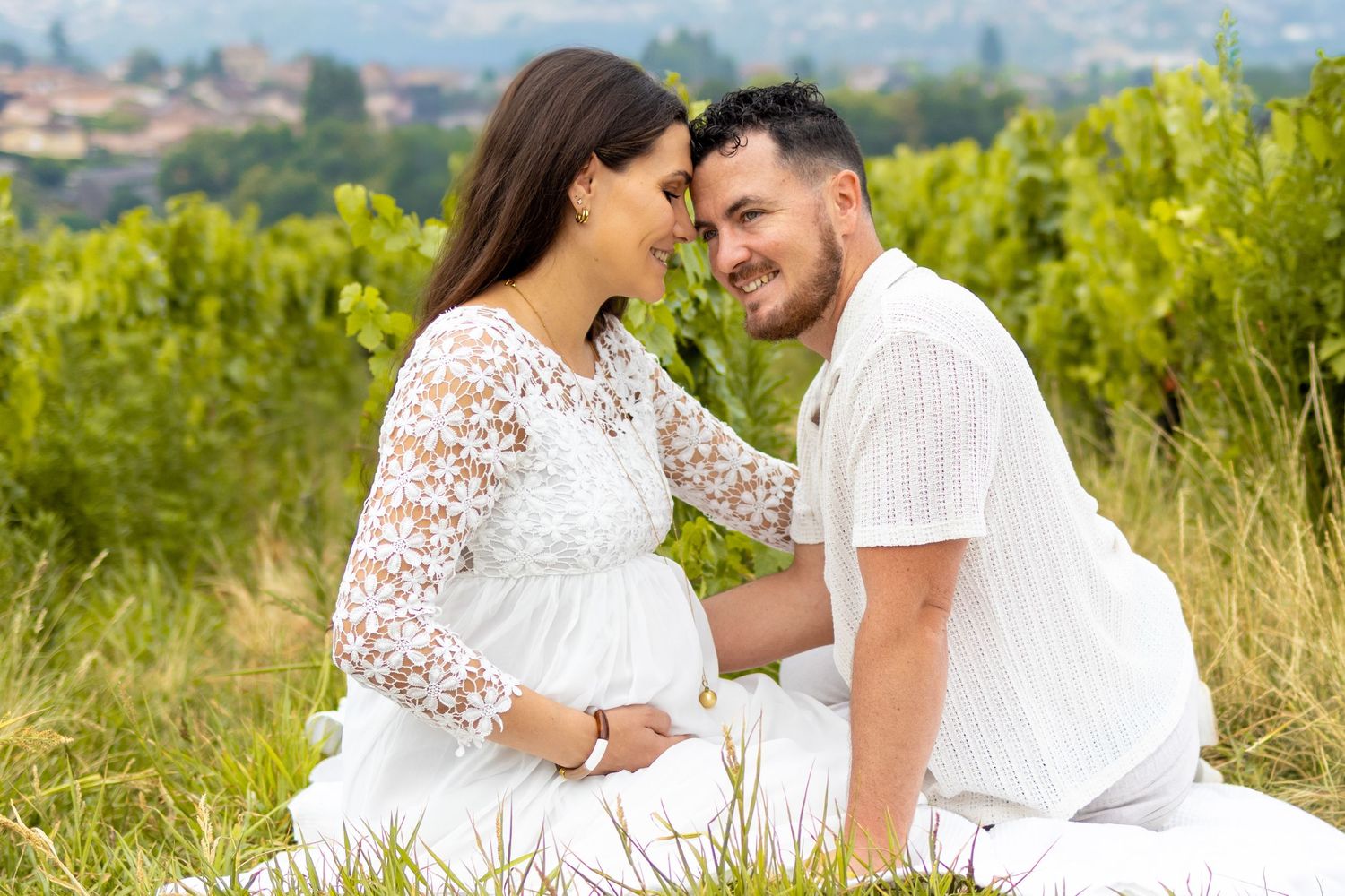 Pregnant couple in white outfits sharing a tender moment in a lush vineyard setting outdoors.