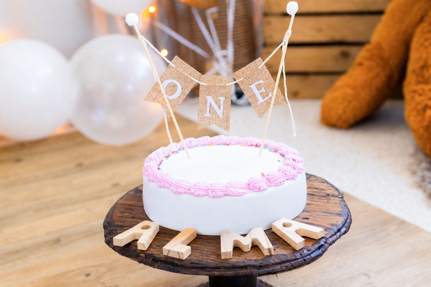 White first birthday cake with pink frosting, ONE banner topper, and wooden letter name on a rustic cake stand.