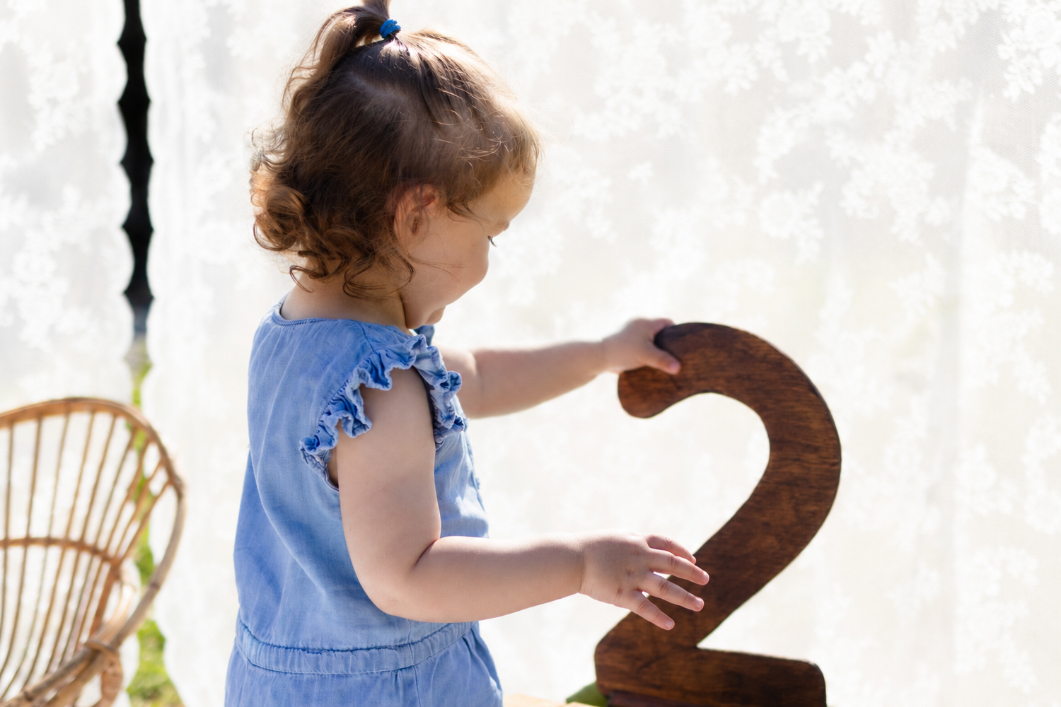 Toddler girl in blue dress holding a wooden number 2, celebrating her second birthday outdoors.