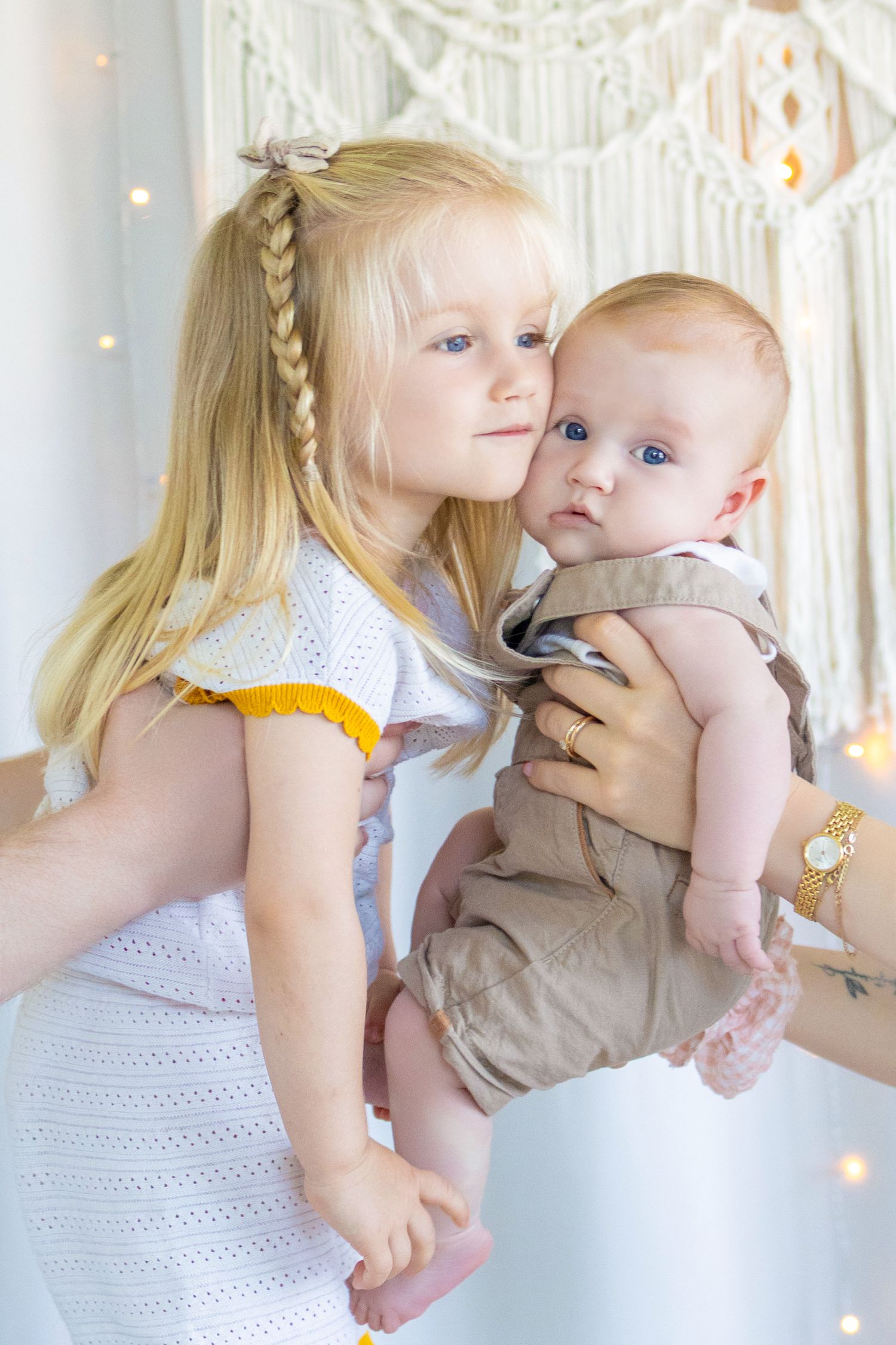 Blonde girl in white dress hugging a blue-eyed baby in tan outfit, held by an adult in a bright room.