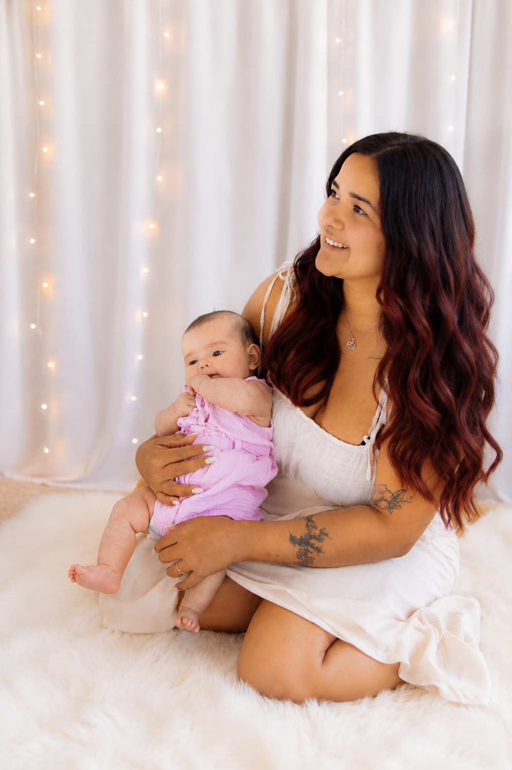 A young woman with long wavy hair holds a baby in a pink outfit, sitting on a white fur rug with fairy lights behind her.