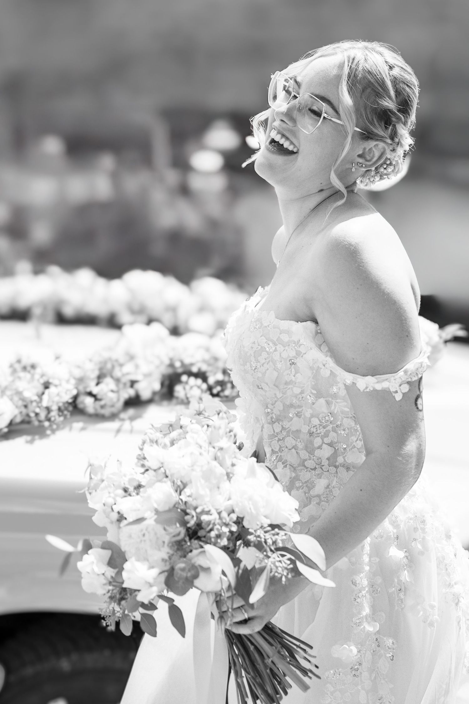 Black and white photo of a laughing bride in a floral gown holding a bouquet beside a flower-adorned car.