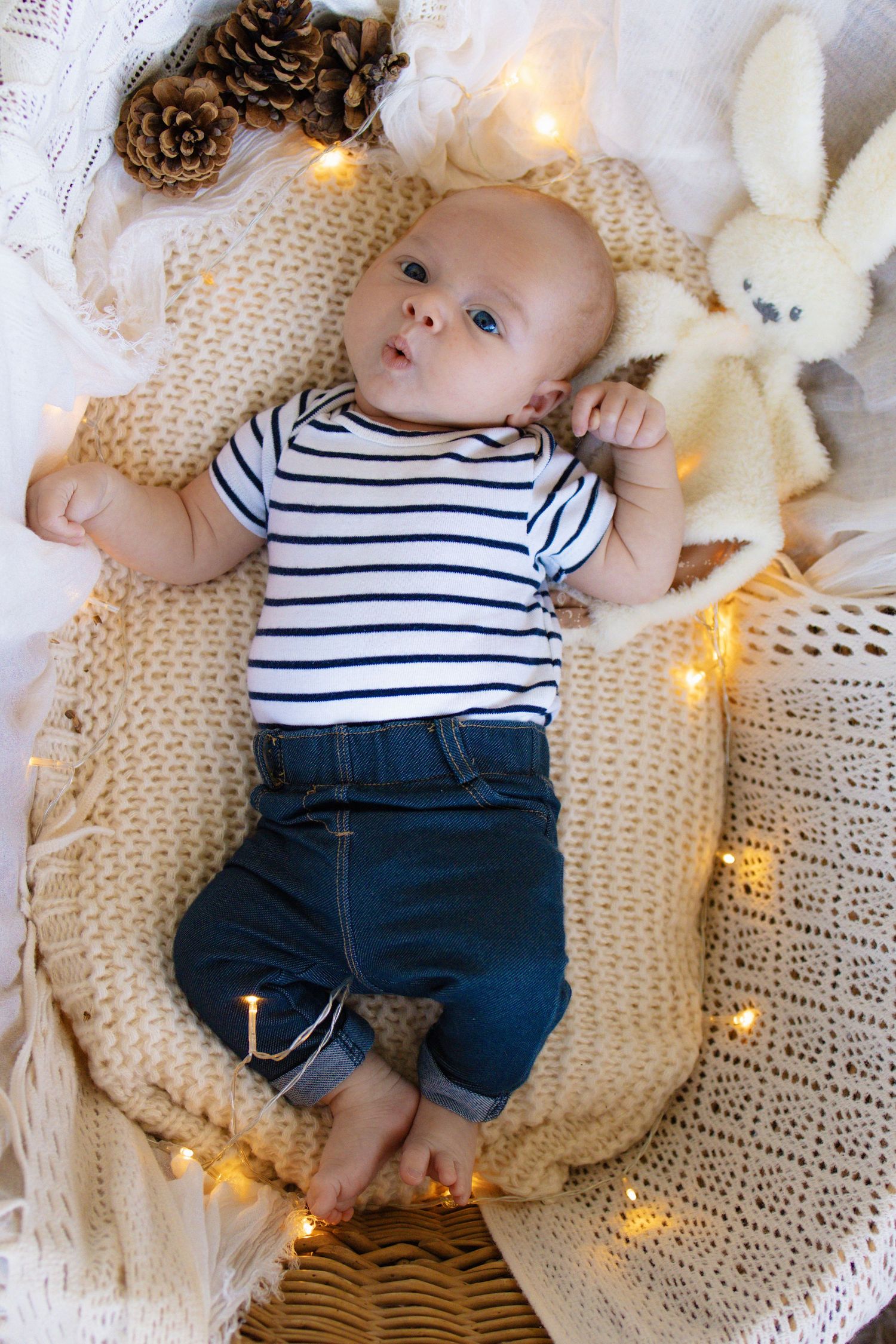 Newborn baby in striped shirt and jeans lying on knit blanket with fairy lights and bunny toy.