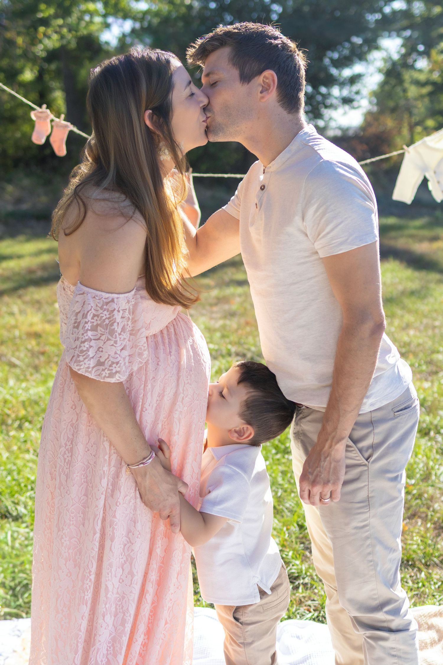 Pregnant woman in pink lace dress kissing man outdoors while young boy hugs her baby bump during maternity photo session.