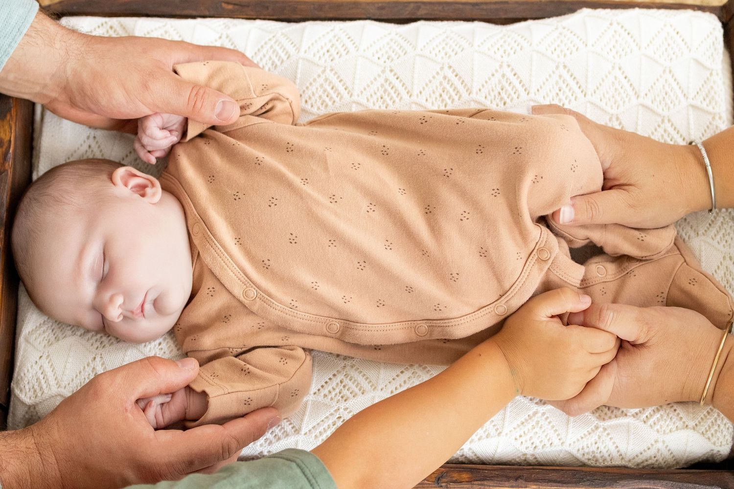 Two adults gently holding a sleeping newborn baby dressed in a brown onesie on a changing pad.