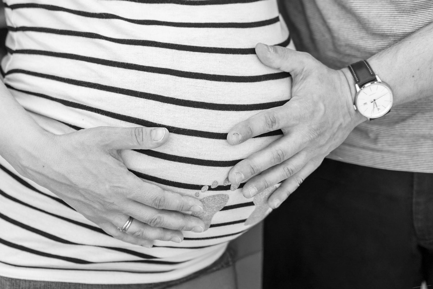 Black and white photo of pregnant woman in striped shirt with partner's hands cradling baby bump.