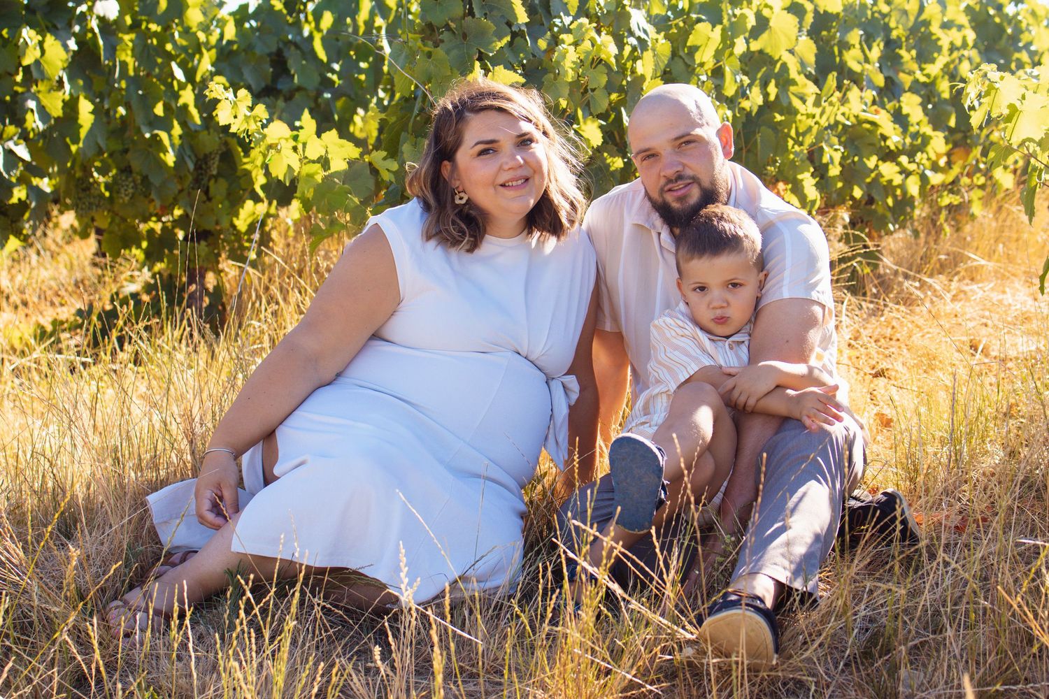 Pregnant woman in white dress sits with husband and young son in golden vineyard field during family maternity photo session.