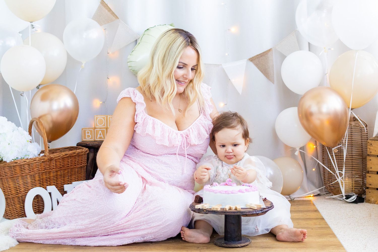 A pregnant woman in a pink dress sits with her baby celebrating a first birthday cake smash with balloons.