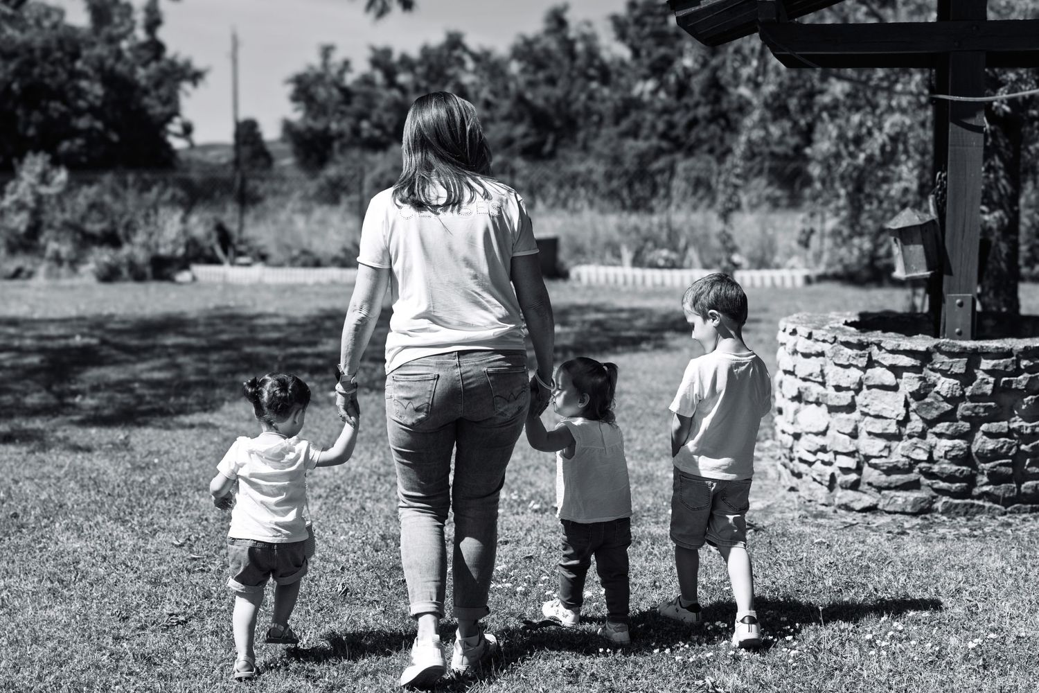 Black and white photo of a woman holding hands with three young children walking through a grassy park near a stone well.