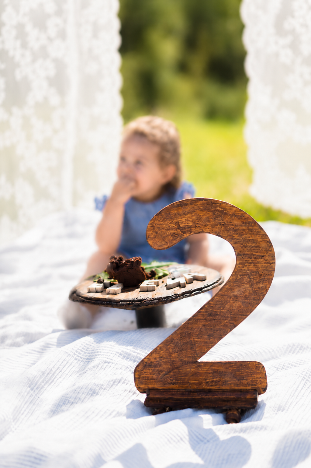 Wooden number 2 sign in foreground with blurred toddler eating cake on white blanket outdoors.