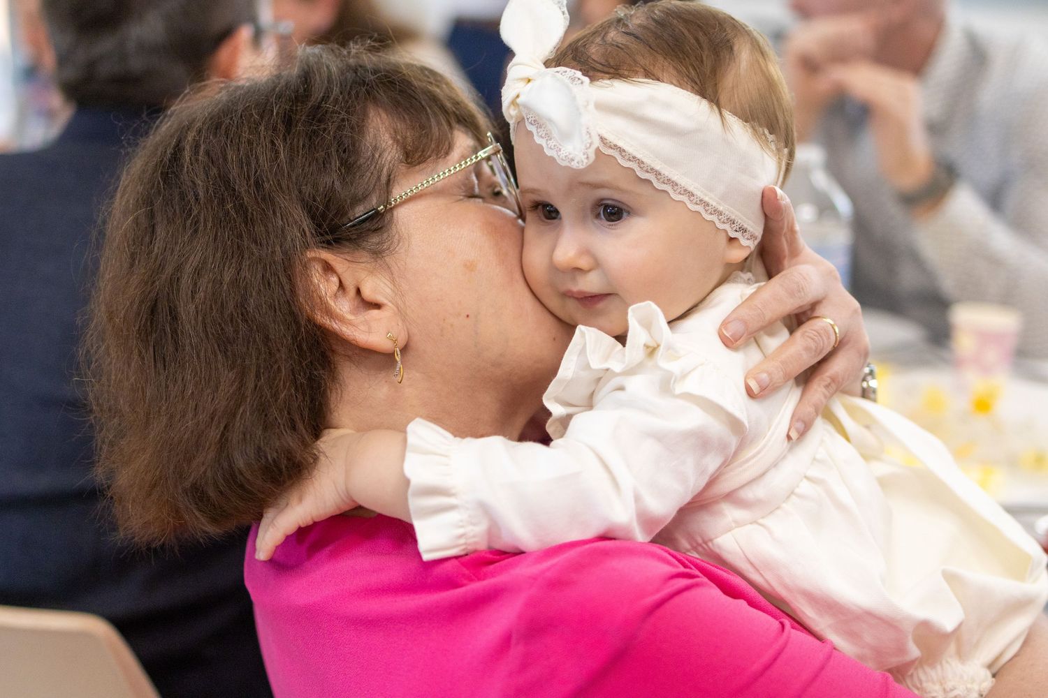 Grandmother kissing a baby girl wearing a white bow headband at a family gathering.