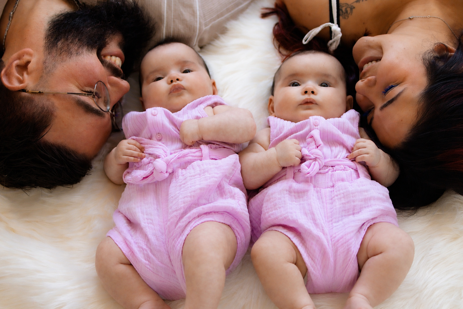 Two adults lying beside twin baby girls dressed in matching pink outfits on a soft white fluffy blanket.
