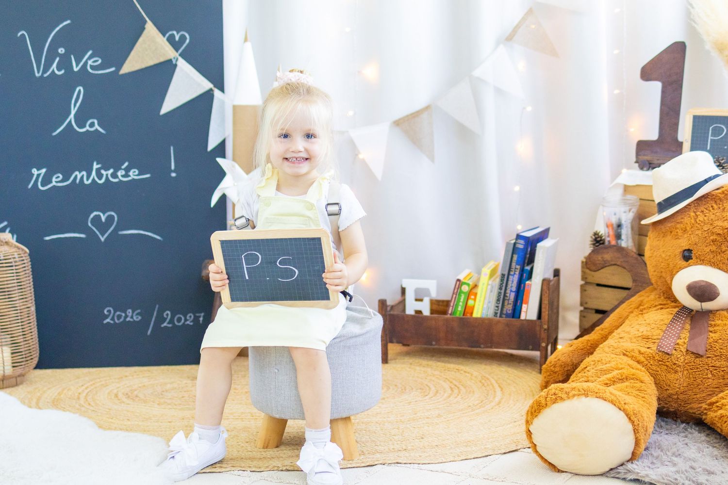 Smiling toddler girl holding a chalkboard sign in a decorated classroom with a teddy bear and back-to-school decor.