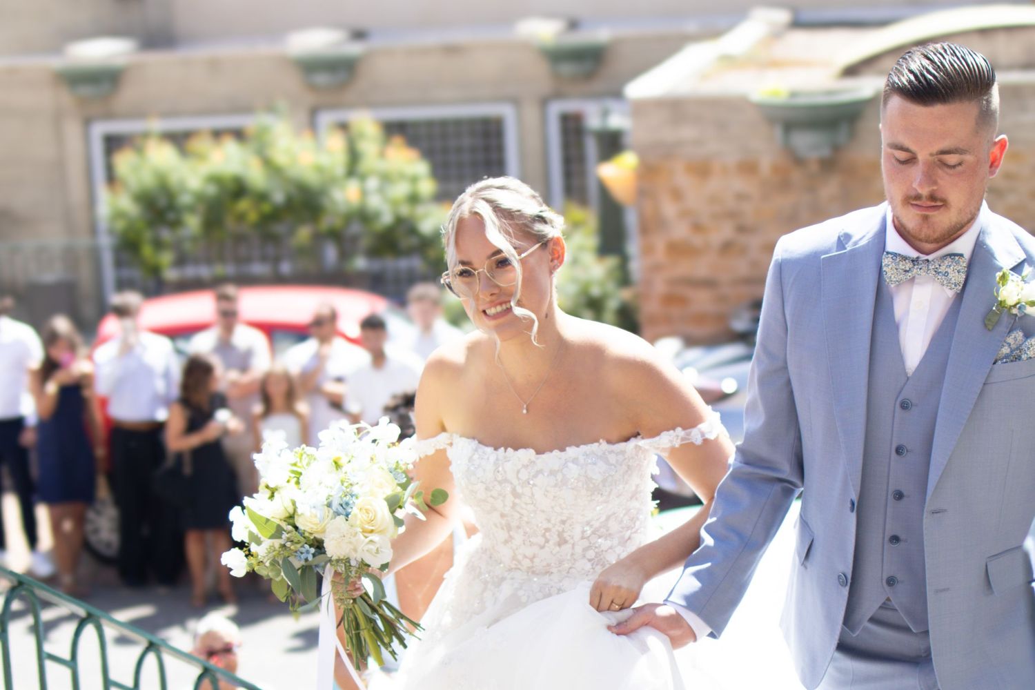 Smiling bride in off-shoulder lace gown holds white bouquet beside groom in gray suit with bow tie at outdoor wedding.
