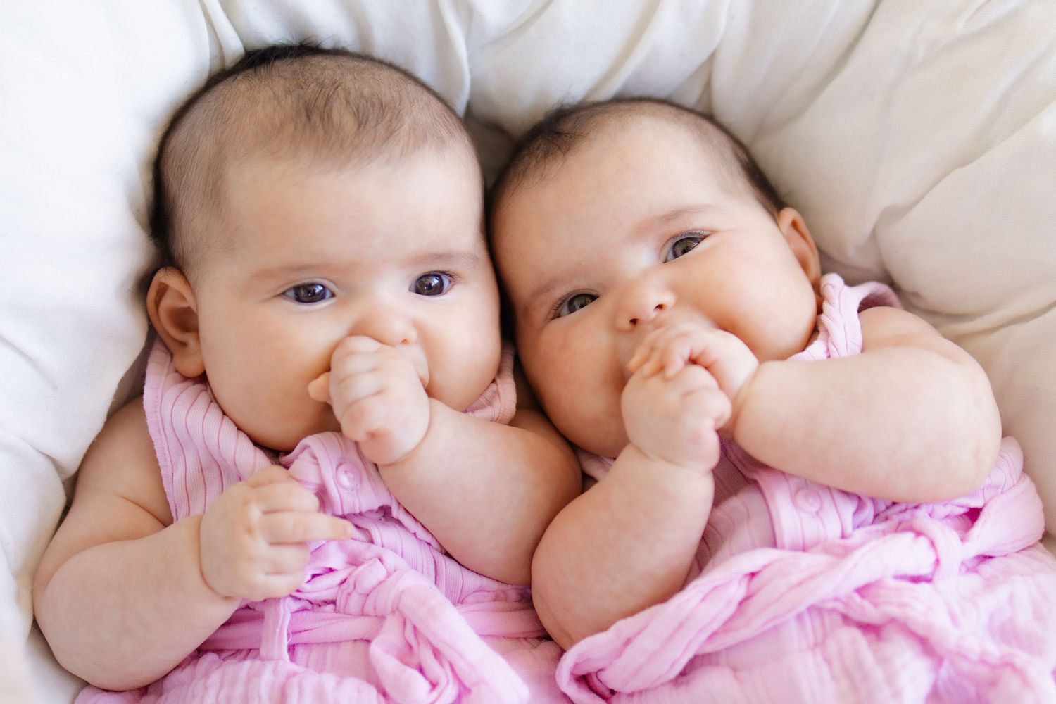 Two adorable twin babies in pink outfits lying together on a white blanket, sucking their fists.