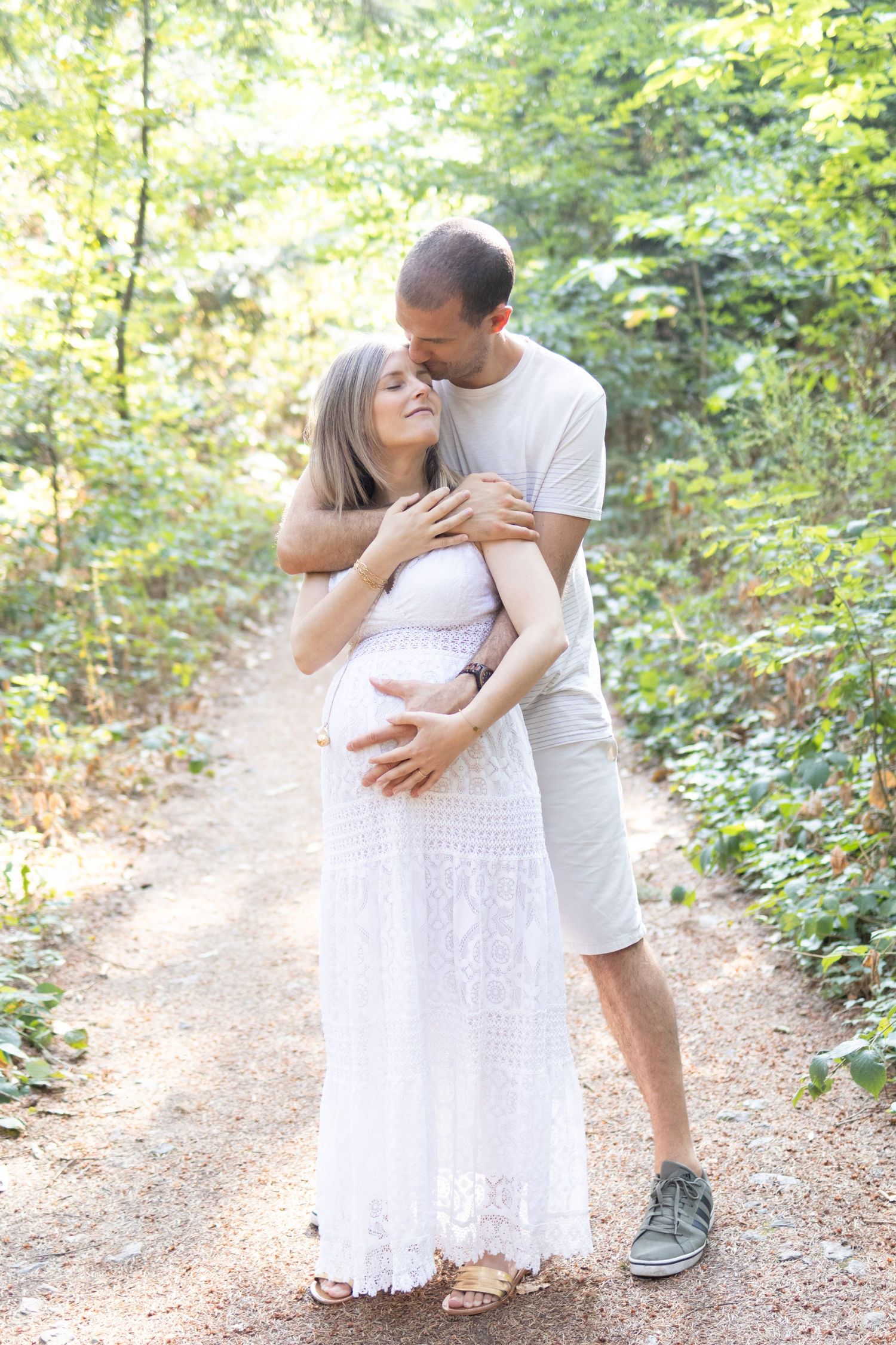 Pregnant couple embracing on a forest path, woman in white lace dress, man kissing her head while holding her baby bump.