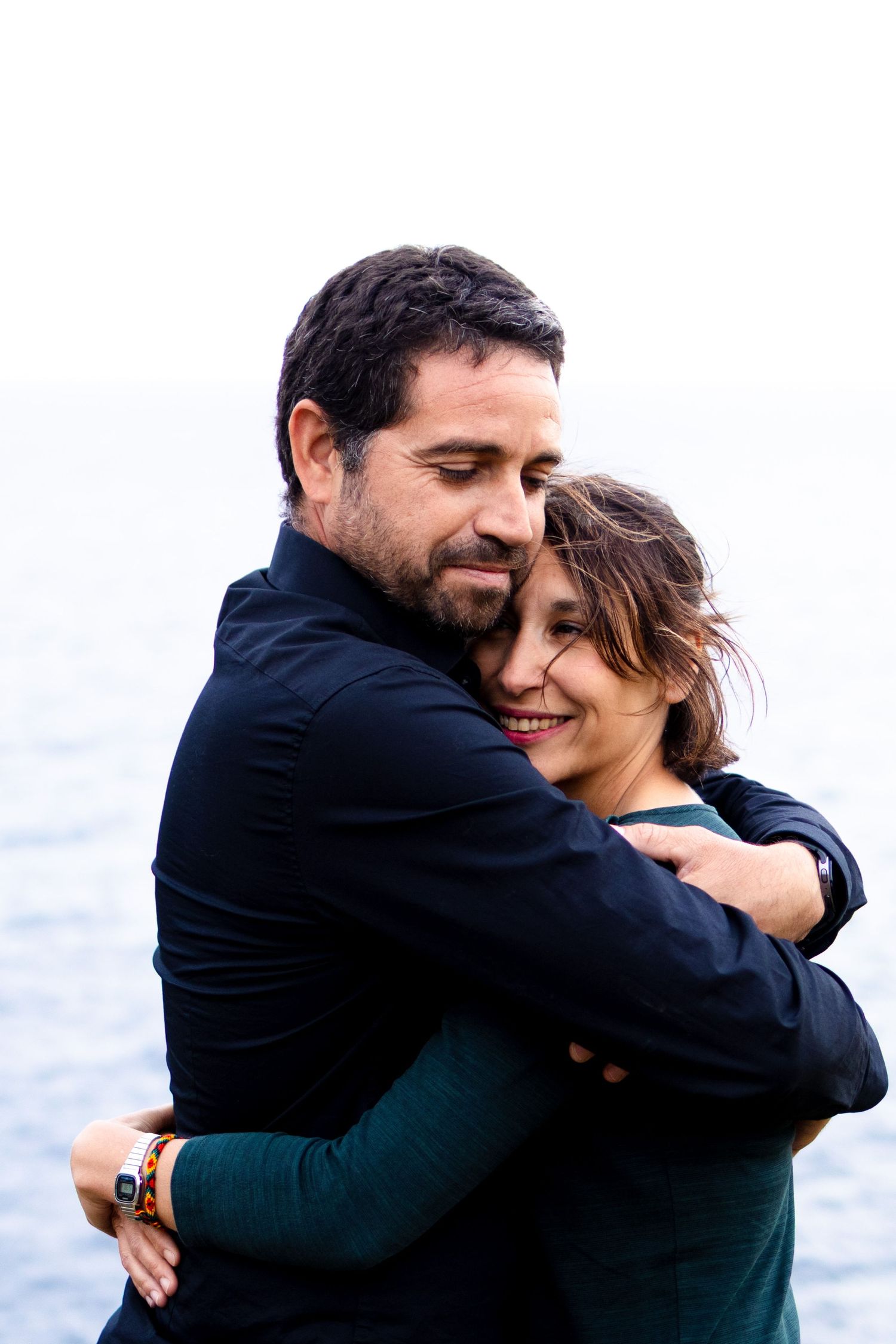 Loving couple embracing warmly by the sea, smiling with eyes closed in a heartfelt hug.