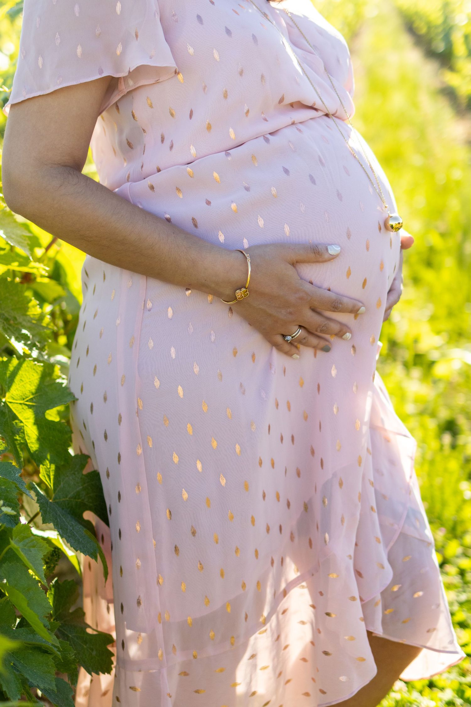 Pregnant woman in pink polka dot dress cradling baby bump outdoors in sunlit green garden.