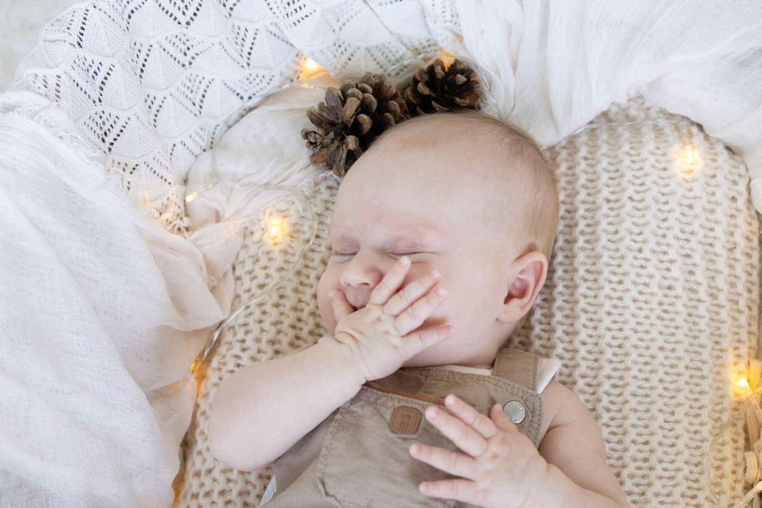 Sleeping baby with pine cone hair accessories surrounded by warm fairy lights on a cozy knit blanket.