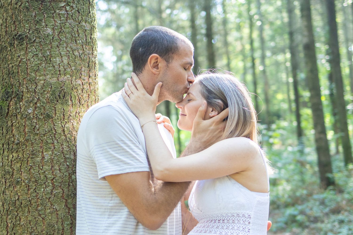 Couple sharing a tender kiss leaning against a tree in a lush green forest, dressed in white outfits.