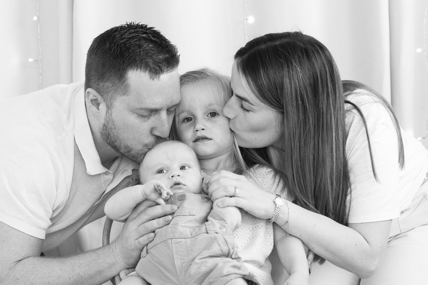 Black and white family portrait of parents kissing toddler on cheeks while holding newborn baby.