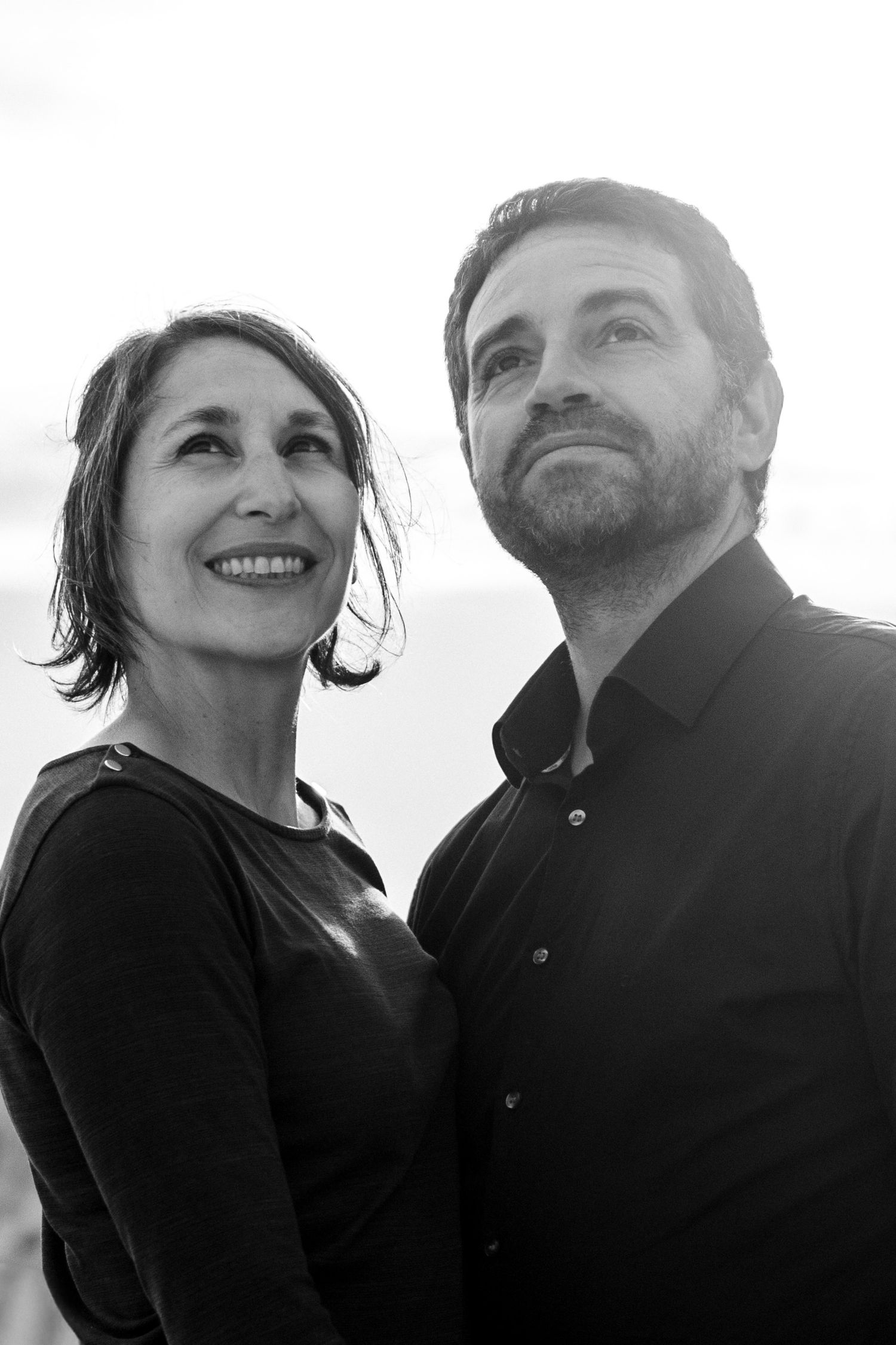 Black and white portrait of a smiling middle-aged couple looking up, dressed in dark clothing outdoors.