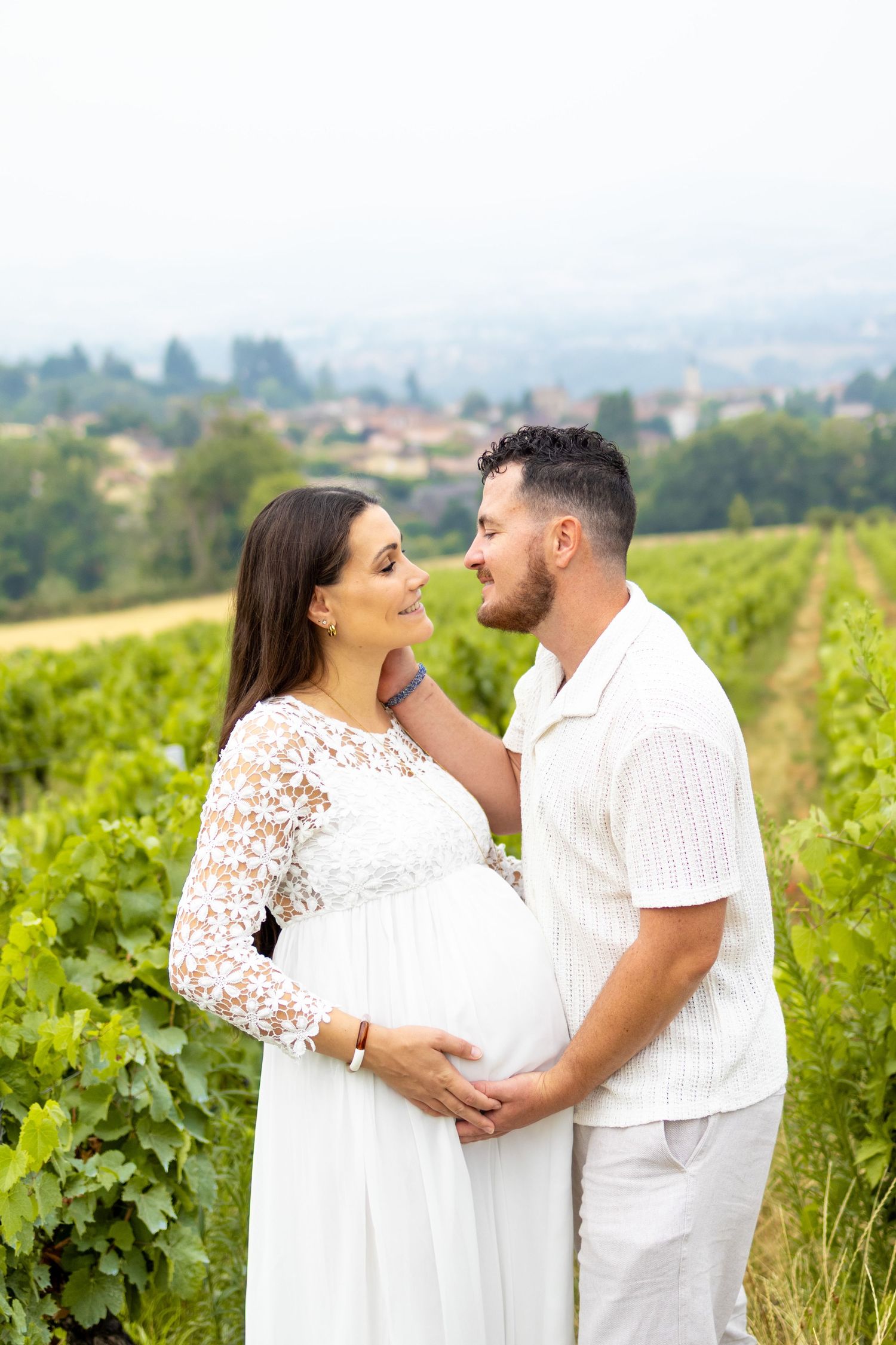 Couple in white outfits sharing a tender moment in a vineyard, woman in lace dress cradling pregnant belly.